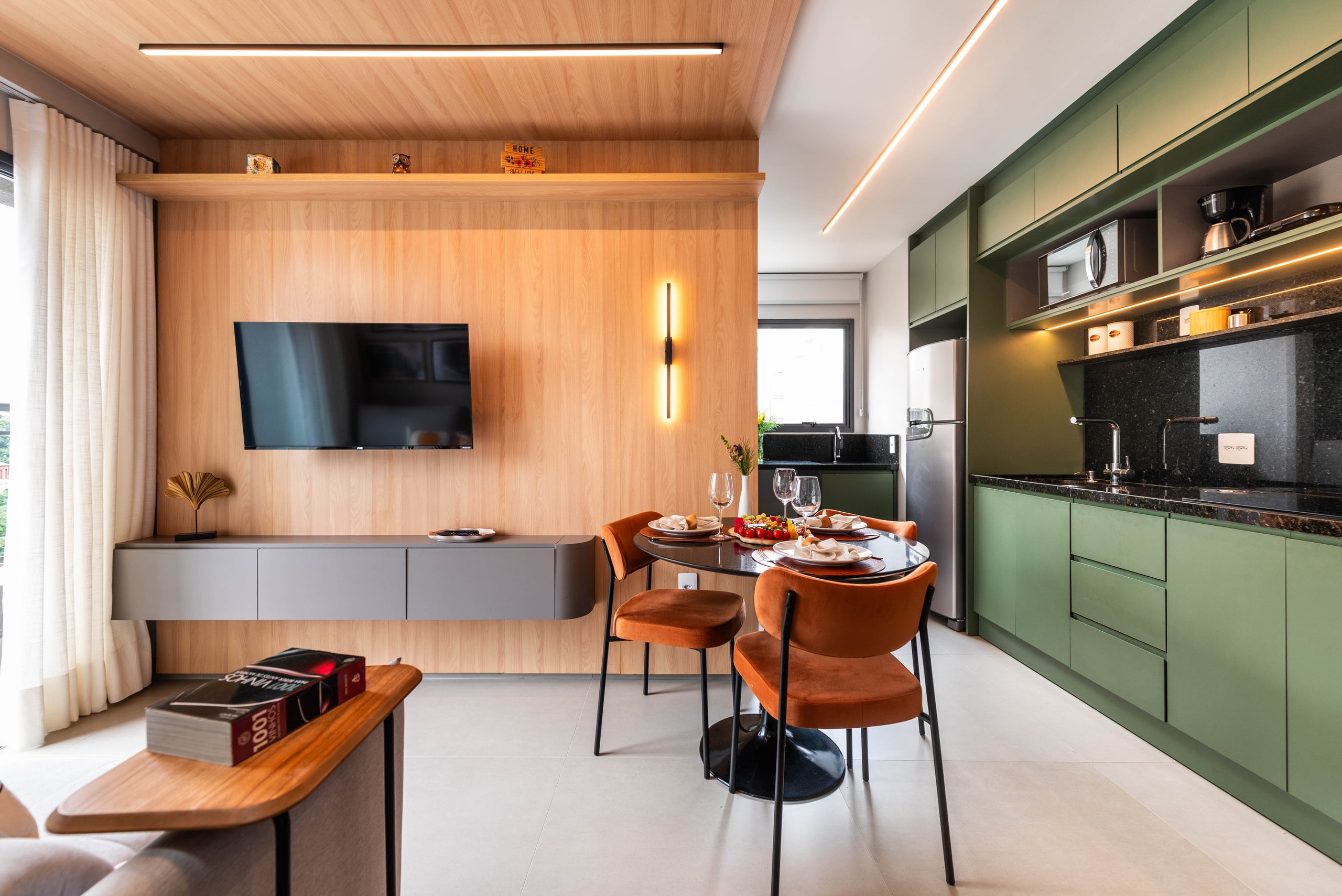 Sala de estar e jantar integradas à cozinha verde, com painel de madeira, mesa redonda posta e duas cadeiras de veludo terracota